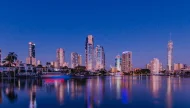 A stunning photograph of the Gold Coast, Australia skyline at dusk, with the illuminated skyscrapers of Surfers Paradise, including the iconic Q1 tower, reflecting in the calm water.