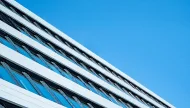 A dynamic low-angle, diagonal shot of a modern architectural facade, showing a repeating pattern of white horizontal lines and angled glass windows against a clear, vibrant blue sky.