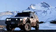 A heavily modified, mud-covered white Jeep Grand Cherokee equipped for off-roading, parked in a rugged landscape with a large, snow-capped mountain in the background.