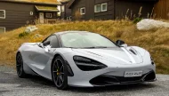 A silver McLaren 720S supercar with white racing stripes and a wedding bow on the mirror, parked in front of traditional Norwegian houses with green sod roofs.
