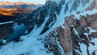 A spectacular aerial view of a snow-covered mountain range at sunrise, with a steep, rocky cliff in the foreground and a dark blue alpine lake nestled in the valley below.