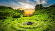 A vibrant, magical sunset over the Fairy Glen on the Isle of Skye, Scotland, featuring lush green hills, the iconic Castle Ewen rock formation, and a stone labyrinth in the foreground.