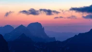 A serene view of layered blue mountain silhouettes fading into the distance under a beautiful pastel pink, orange, and blue sunset sky.