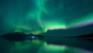 A spectacular display of the green and purple Aurora Borealis (Northern Lights) dancing in a star-filled sky, perfectly reflecting over the calm water of a lake in front of a mountain silhouette.
