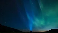 Silhouette of a person shining a headlamp up into a star-filled sky with the green and blue Aurora Borealis (Northern Lights) overhead in a dark valley.